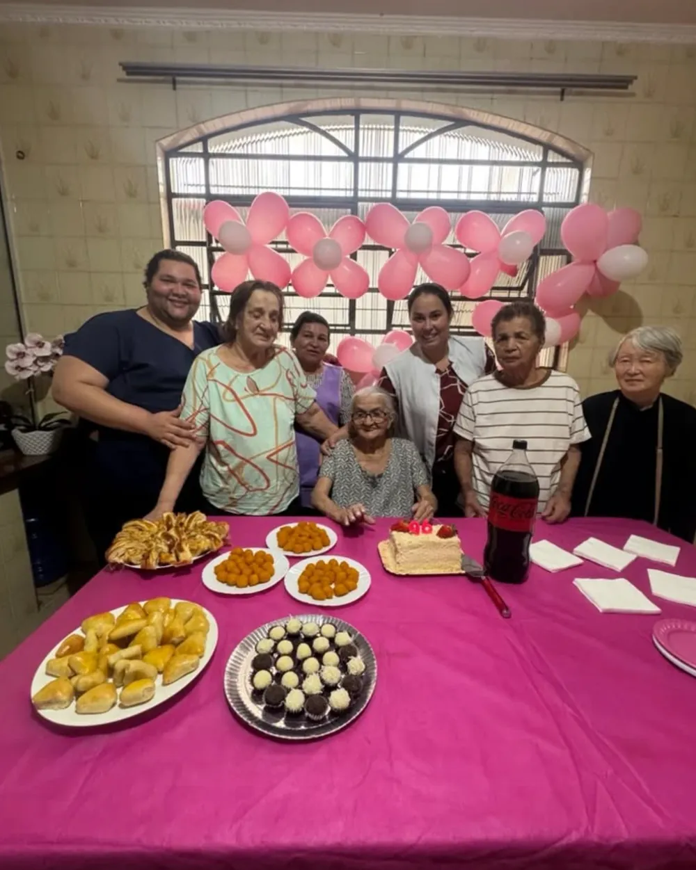 Residentes e cuidadoras reunidas em volta da mesa de festa, com bolo de 96 anos, salgados, doces e balões rosa na janela