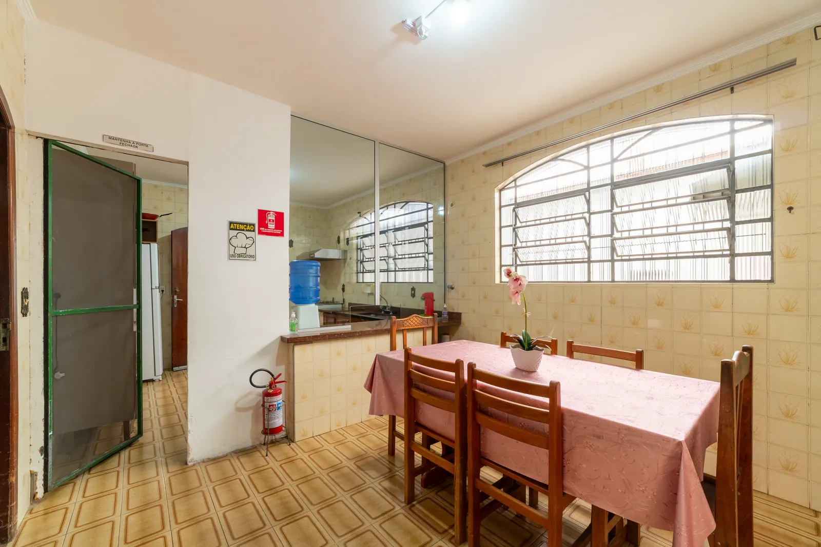 Refeitório com mesa de toalha rosa, cadeiras de madeira e vaso de orquídea, com a cozinha ao fundo