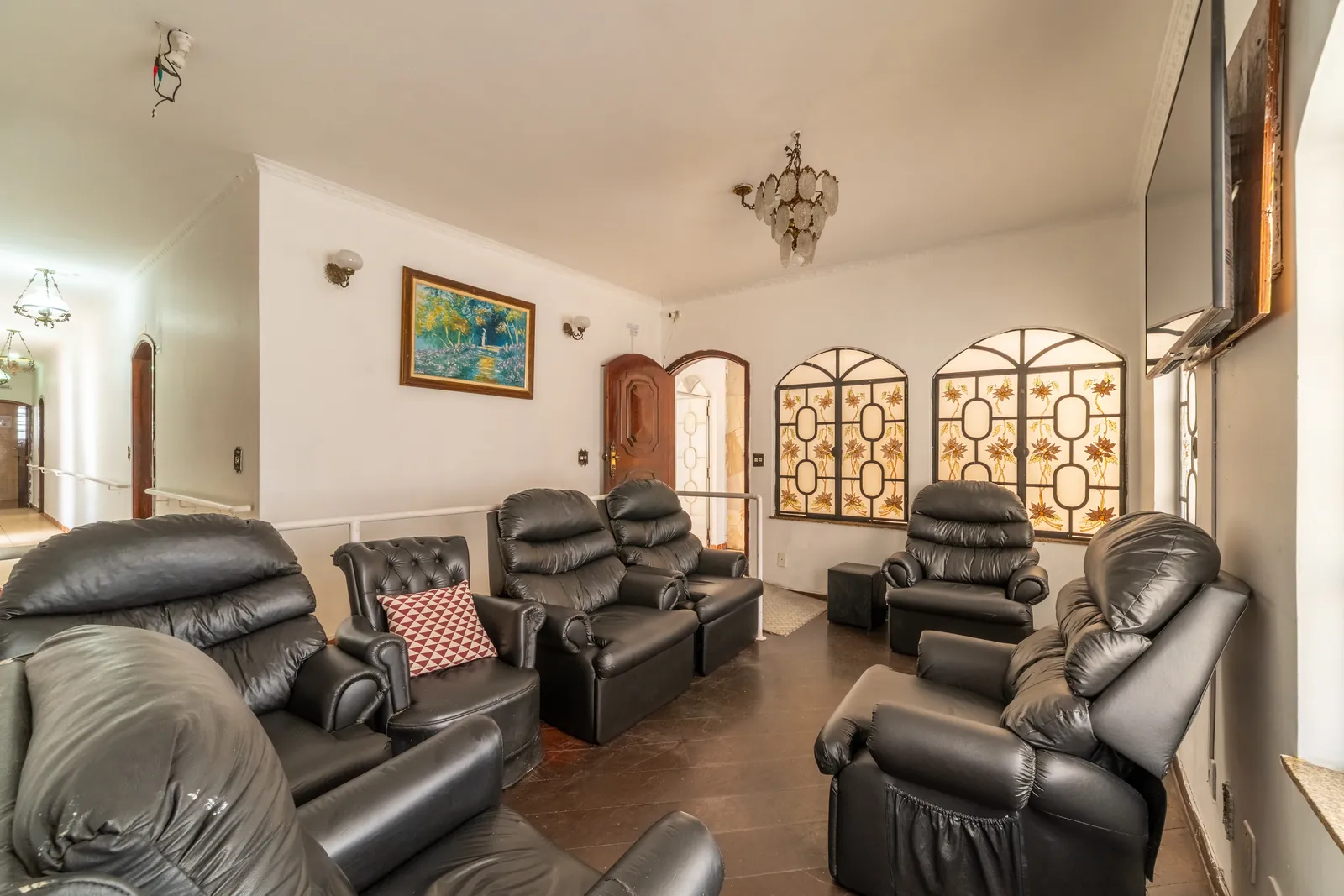 Sala de estar com poltronas reclináveis de couro preto, quadro colorido na parede, lustre e janelas em arco com vitrais