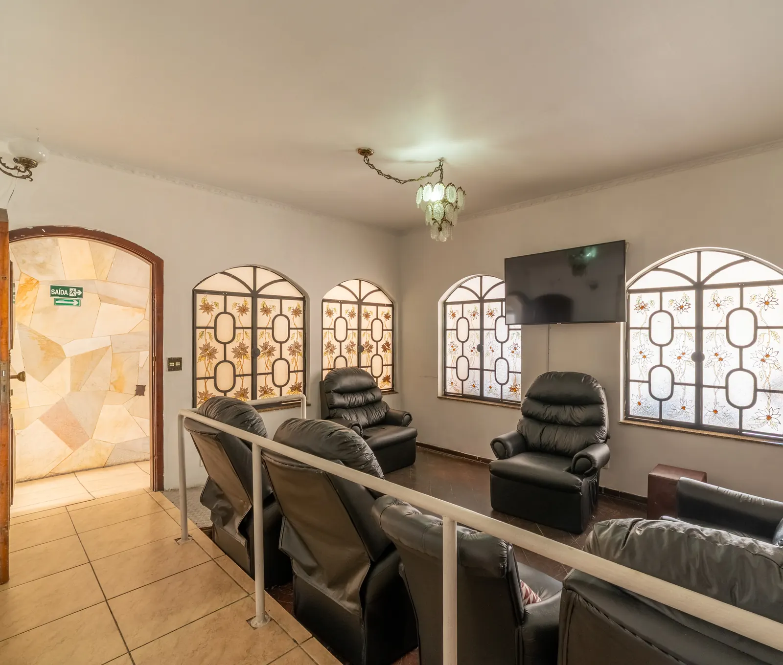 Sala de estar com poltronas reclináveis de couro, televisão e janelas em arco com vitrais